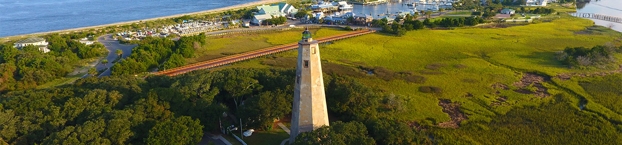 An aerial view of a lighthouse with a marina behind it in the distance.